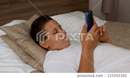 Teenage boy lying in bed, looking at smartphone screen and smiling.  133203302