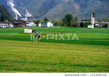 A tractor is engaged in harvesting on a small private agricultural field against the backdrop of the Alpine mountains in Austria 133204338