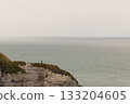 Lone figure standing on a high coastal cliff overlooking a calm, expansive sea under a soft overcast sky. 133204605