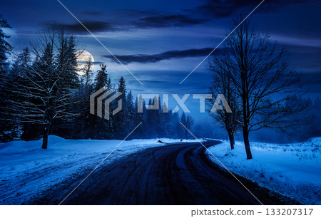 winding road through winter forest at night. beautiful nature scenery with naked trees on foggy weather in full moon light. transportation background. backdrop for fiction art or mystery concepts winding road through winter forest at night. beautiful nature scenery with naked trees on foggy weather in full moon light. transportation background. backdrop for fiction art or mystery concepts 133207317