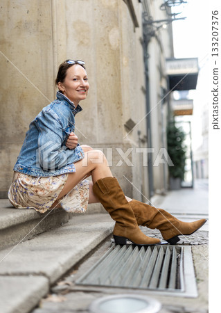 Portrait of young adult woman wear floral dress denim jacket brown suede boots  sitting on stairs outdoors in old European city. Stylish female person enjoy walk travel urban lifestyle summertime day 133207376