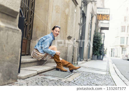 Portrait of young adult woman wear floral dress denim jacket brown suede boots sitting on stairs outdoors in old European city. Stylish female person enjoy walk travel urban lifestyle summertime day Portrait of young adult woman wear floral dress denim jacket brown suede boots sitting on stairs outdoors in old European city. Stylish female person enjoy walk travel urban lifestyle summertime day 133207377