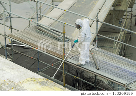 Worker in protective suit cleans scaffolding at construction site near a river in daylight while ensuring safety and cleanliness during maintenance work 133209199