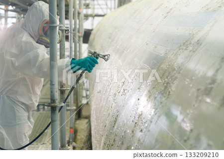 Worker in protective gear spray painting a large industrial tank at a construction site during daylight hours Worker in protective gear spray painting a large industrial tank at a construction site during daylight hours 133209210