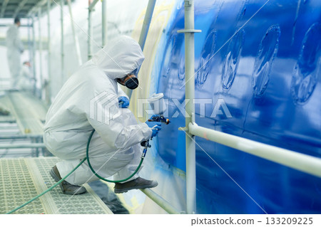 Technician applies blue paint to aircraft fuselage in maintenance facility during daylight hours Technician applies blue paint to aircraft fuselage in maintenance facility during daylight hours 133209225