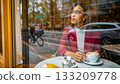 Thoughtful woman enjoying coffee in a city cafe. Woman seated in a city cafe, holding a cup of coffee. She appears thoughtful, looking out the window.  133209778