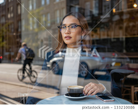 Thoughtful woman enjoying coffee in a cafe with city reflections. A woman sits at a table in a cafe, her hand gently holding a cup of coffee. Thoughtful woman enjoying coffee in a cafe with city reflections. A woman sits at a table in a cafe, her hand gently holding a cup of coffee. 133210086