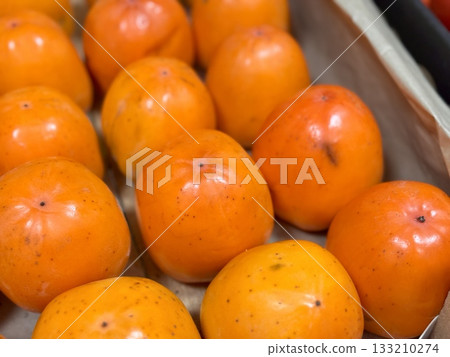 A pile of ripe juicy orange persimmons in a box, on sale at a supermarket vegetable stand, showcases organic, vegetarian and healthy food. Close-up. 133210274