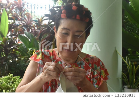 Asian elder woman sewing cloth for repairing in home front yard Asian elder woman sewing cloth for repairing in home front yard 133210935