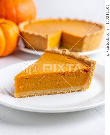 A single, smooth pumpkin pie slice sits neatly on a pristine white plate, waiting for the first spoonful. A single, smooth pumpkin pie slice sits neatly on a pristine white plate, waiting for the first spoonful. 133211102