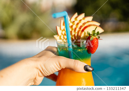 A hand holds a colorful cocktail featuring layers of orange, blue, and green, garnished with apple slices and a strawberry. The setting is a sunny poolside during summer A hand holds a colorful cocktail featuring layers of orange, blue, and green, garnished with apple slices and a strawberry. The setting is a sunny poolside during summer 133211347