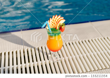 A vibrant tropical drink sits on the edge of a pool, adorned with sliced fruit and a colorful straw. Sunlight reflects on the water, creating a refreshing summer vibe A vibrant tropical drink sits on the edge of a pool, adorned with sliced fruit and a colorful straw. Sunlight reflects on the water, creating a refreshing summer vibe 133211354