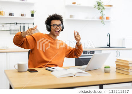 A joyful man with headphones points at the camera while working on a laptop at home. A joyful man with headphones points at the camera while working on a laptop at home. 133211357