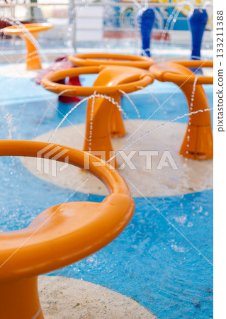 Children can enjoy playtime in a lively splash pad featuring colorful water structures. Bright orange fountains spray water, creating a playful atmosphere on a sunny day 133211388