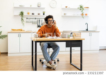 A young man with headphones smiles while working on a laptop at a table in his kitchen. A young man with headphones smiles while working on a laptop at a table in his kitchen. 133211397
