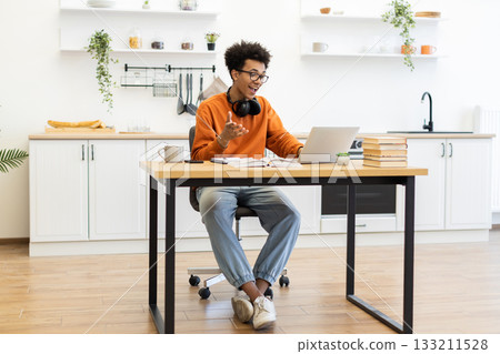 A young man with glasses is working on his laptop at a table in a modern kitchen setting. 133211528
