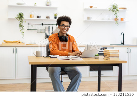 A young man with glasses smiles while working on his laptop at a wooden table in a bright kitchen setting. 133211530