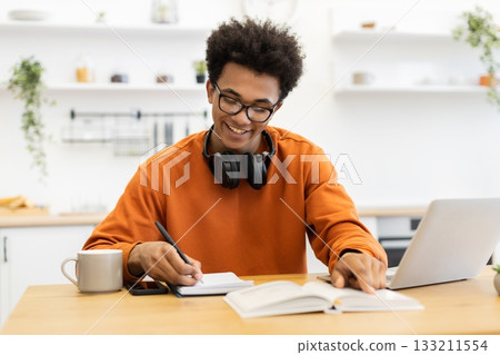 A young Black man smiles while studying at a table with a laptop, notebook, and book. 133211554