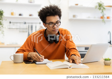 A young man with glasses is studying at home, using a laptop and taking notes. A young man with glasses is studying at home, using a laptop and taking notes. 133211564