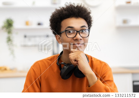 A young Black man with glasses smiles, resting his chin on his hand, with headphones around his neck. A young Black man with glasses smiles, resting his chin on his hand, with headphones around his neck. 133211572