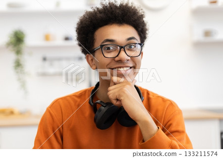 A cheerful young man with glasses smiles while resting his chin on his hand, with headphones around his neck. 133211574