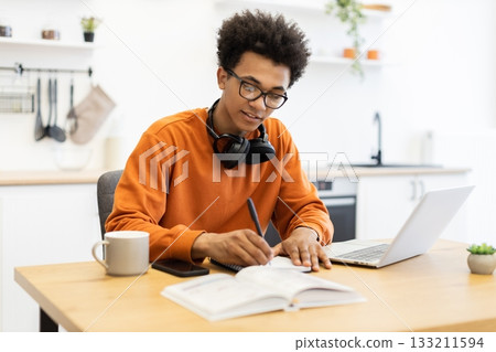 A young Black man wearing glasses and headphones studies at a table with a laptop and notebook in a bright kitchen setting. A young Black man wearing glasses and headphones studies at a table with a laptop and notebook in a bright kitchen setting. 133211594