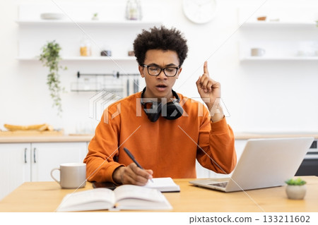 A young man with glasses is working on his laptop and writing in a notebook while having an idea. A young man with glasses is working on his laptop and writing in a notebook while having an idea. 133211602