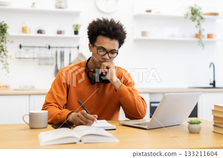 A young Black man wearing glasses is studying at a table, with a laptop, notebook, and coffee cup. A young Black man wearing glasses is studying at a table, with a laptop, notebook, and coffee cup. 133211604