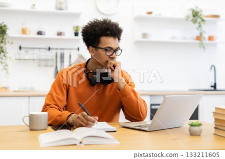 A young man with glasses is working on a laptop at home, taking notes and looking thoughtful. 133211605