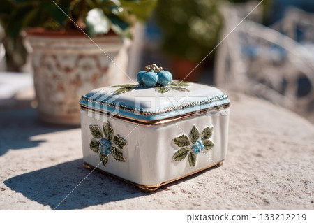 Elegant ceramic box with blueberry and leaf details standing on a sunlit outdoor table. Perfect for holiday d cor, gifts, summer settings, and vintage-inspired designs. Elegant ceramic box with blueberry and leaf details standing on a sunlit outdoor table. Perfect for holiday d cor, gifts, summer settings, and vintage-inspired designs. 133212219