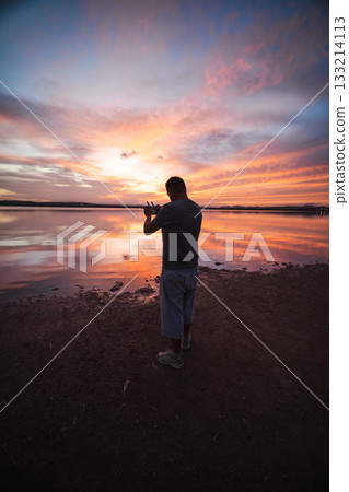 Man standing by the lake at sunset, controlling a drone under a colorful orange and purple sky reflected in calm water. Man standing by the lake at sunset, controlling a drone under a colorful orange and purple sky reflected in calm water. 133214113