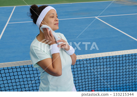 Confident Woman Athlete On Blue Tennis Court After Workout Break With Towel And Headband Confident Woman Athlete On Blue Tennis Court After Workout Break With Towel And Headband 133215344