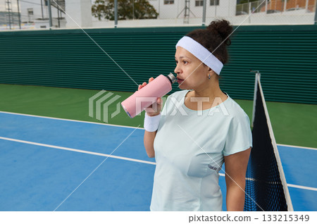 Athletic Woman Drinking Water On Tennis Court Between Games During Workout Session 133215349
