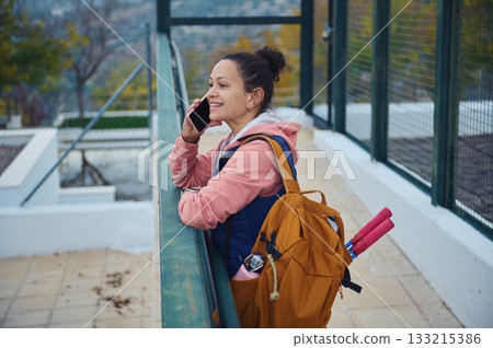 Smiling Woman With Backpack Talks On Phone At Outdoor Sports Court, Ready For Exercise Smiling Woman With Backpack Talks On Phone At Outdoor Sports Court, Ready For Exercise 133215386