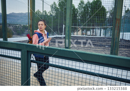 Woman With Backpack Walks Past Fence and Uses Phone in Outdoor Park Setting Woman With Backpack Walks Past Fence and Uses Phone in Outdoor Park Setting 133215388