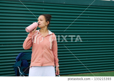 Woman in Pink Hoodie Drinking Water Bottle Outdoors By Green Wall During Workout Break 133215393