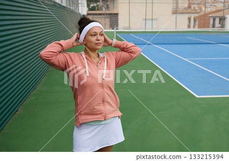 Woman Athlete Stretches On Tennis Court Between Green Fence And Blue Courts 133215394