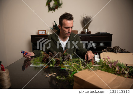 Man Crafts Christmas Wreaths With Pine Cones And Greenery At Cozy Home Table 133215515