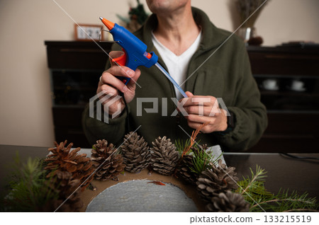 Person Crafting Christmas Wreath With Pinecones And Glue Gun At Home Person Crafting Christmas Wreath With Pinecones And Glue Gun At Home 133215519
