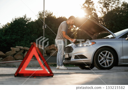 Under the sunlight, red sign, accident, broken automobile, repair. Man is with his car outdoors Under the sunlight, red sign, accident, broken automobile, repair. Man is with his car outdoors 133215582