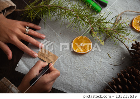Handwriting Christmas Gift Note On Brown Tag With Pine Branch, Orange Slice, And Pinecones 133215639