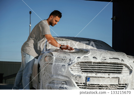 Automotive detailer washing away soap and foam with water high pressure washer. Man is with his car outdoors Automotive detailer washing away soap and foam with water high pressure washer. Man is with his car outdoors 133215670