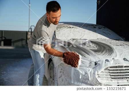 Automotive detailer washing away soap and foam with water high pressure washer. Man is with his car outdoors Automotive detailer washing away soap and foam with water high pressure washer. Man is with his car outdoors 133215671