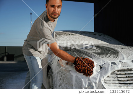 Automotive detailer washing away soap and foam with water high pressure washer. Man is with his car outdoors 133215672