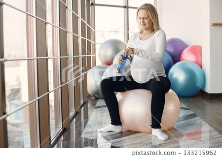 Caucasian pregnant woman in third trimester drinking water from sportive bottle. Woman in bright fitness studio with big windows. Blonde woman wearing whit shirt and black leggins. 133215962