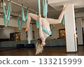 Young woman making antigravity yoga exercises in green hammock at white studio. Professional yoga teacher practicing aerial yoga. Blonde woman wearing beige leggins and shirt. 133215999