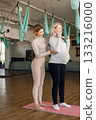 Pregnant woman doing yoga with personal trainer. Yoga trainer assisting pregnant woman while doing exercises with a fitball and dumbbells. Blonde pregnant woman wearing white shirt. 133216000