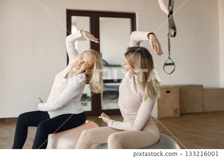 Pregnant woman doing yoga with personal trainer. Yoga trainer assisting pregnant woman while doing exercises with a fitball. Blonde pregnant woman wearing white clothes. Pregnant woman doing yoga with personal trainer. Yoga trainer assisting pregnant woman while doing exercises with a fitball. Blonde pregnant woman wearing white clothes. 133216001