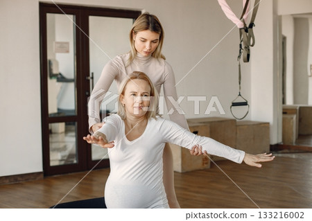 Pregnant woman doing yoga with personal trainer. Yoga trainer assisting pregnant woman while doing exercises. Blonde pregnant woman wearing white clothes. Pregnant woman doing yoga with personal trainer. Yoga trainer assisting pregnant woman while doing exercises. Blonde pregnant woman wearing white clothes. 133216002
