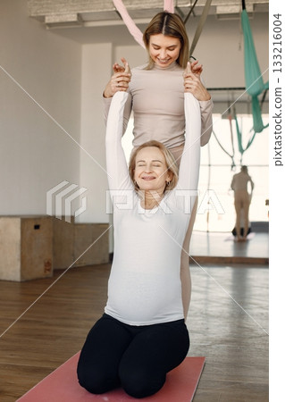 Pregnant woman doing yoga with personal trainer. Yoga trainer assisting pregnant woman while doing exercises. Blonde pregnant woman wearing white clothes. Pregnant woman doing yoga with personal trainer. Yoga trainer assisting pregnant woman while doing exercises. Blonde pregnant woman wearing white clothes. 133216004